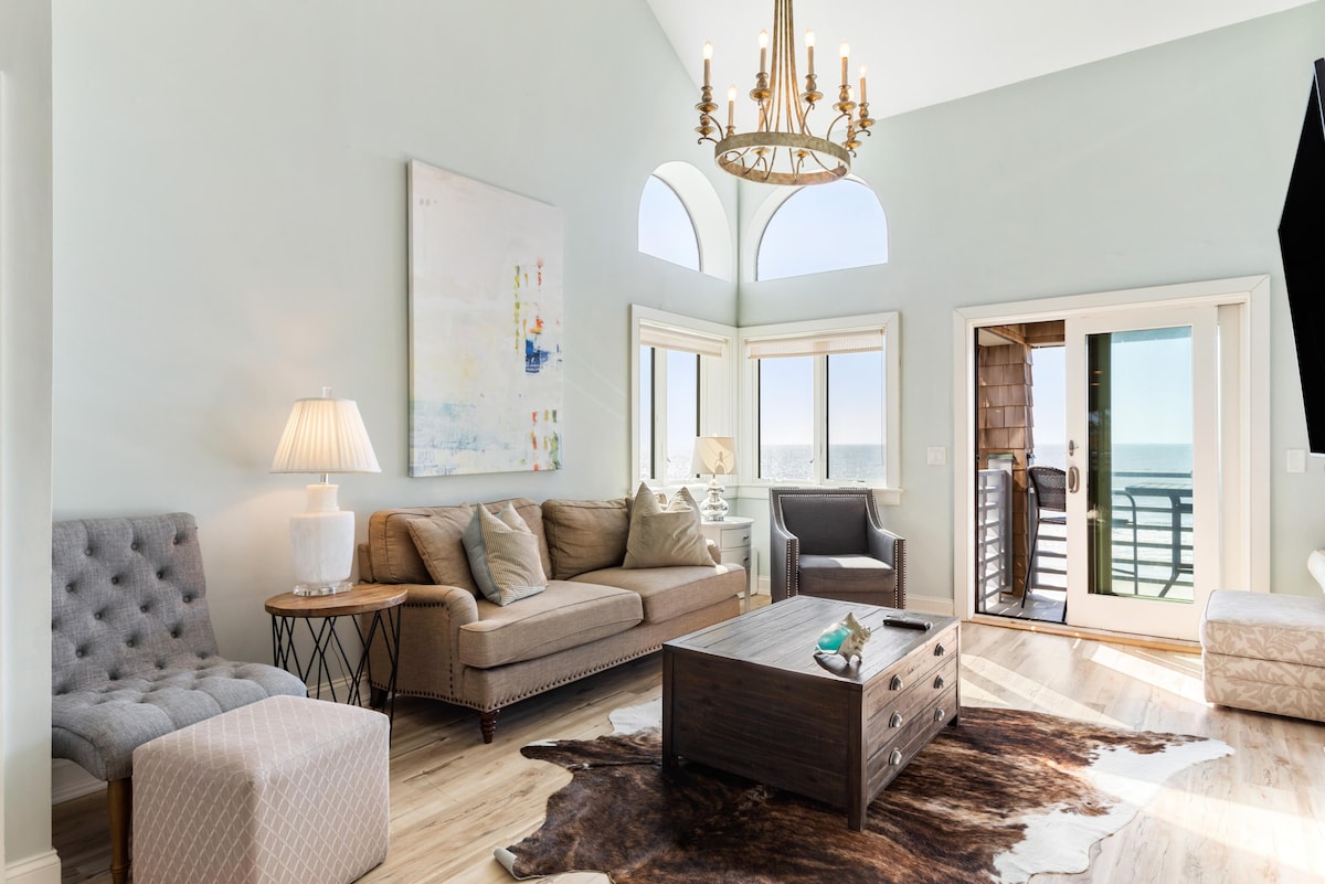 The living room features a vaulted ceiling and large windows that allow ample natural light and stunning ocean views. A neutral-toned sofa and armchair provide comfortable seating, while a wooden coffee table and a light fixture add warmth to the space. Sliding doors lead to an outdoor area.