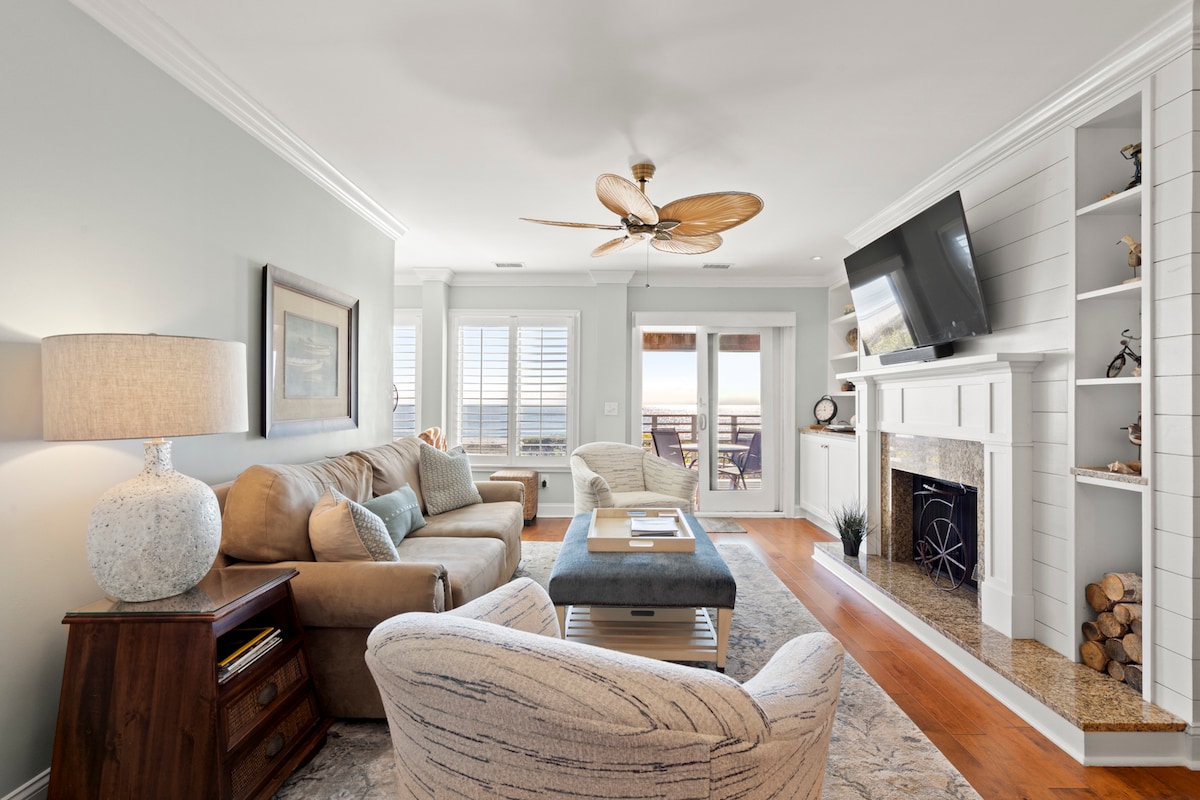 The living area features comfortable seating arrangements, including a beige sofa and a patterned armchair. A flat-screen TV is mounted above a decorative fireplace, flanked by built-in shelves. Natural light floods the space, creating a warm atmosphere, while doors lead to a balcony with ocean views.