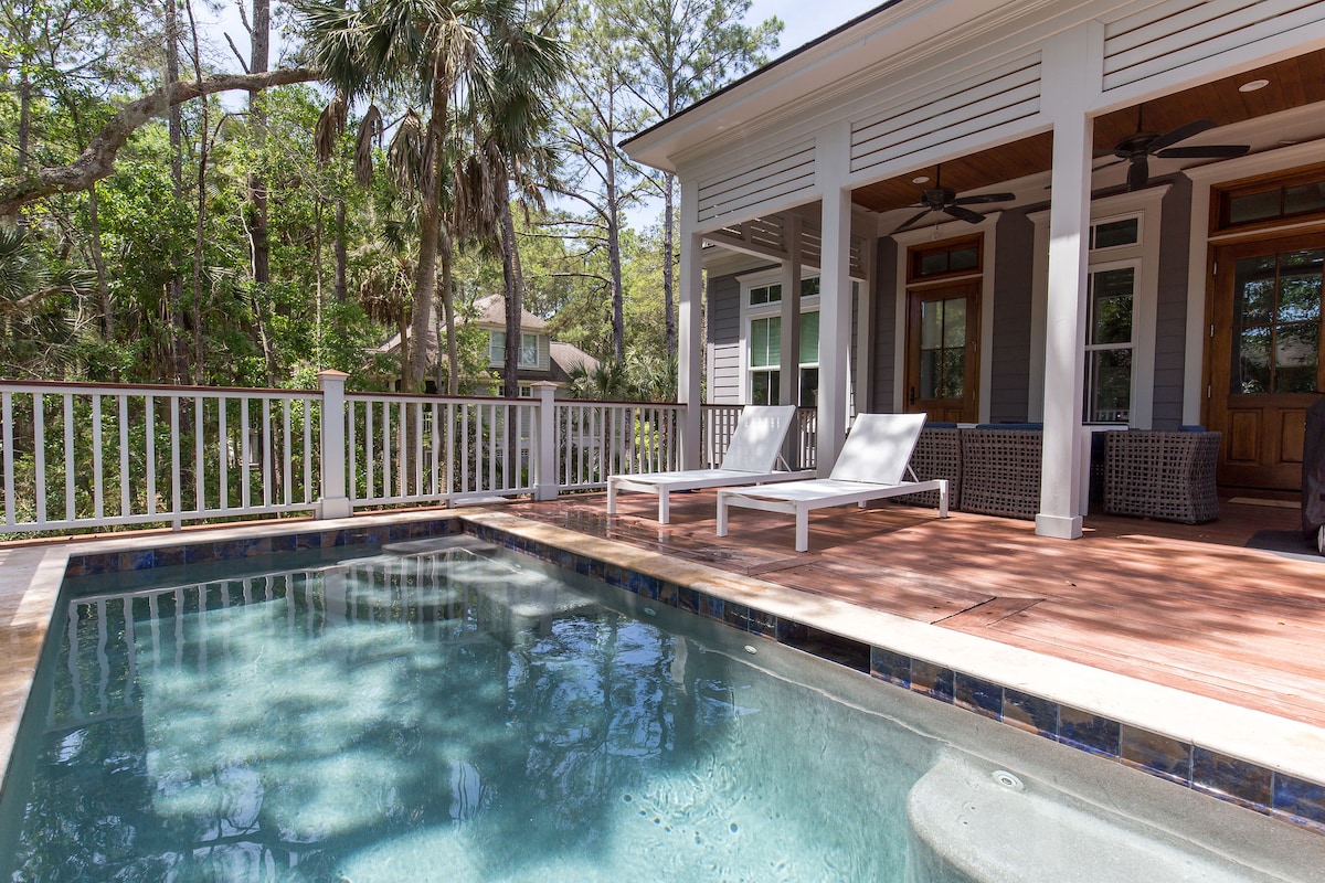 A private pool area is shown with two chaise lounge chairs positioned beside the water. The pool features a textured surface and is surrounded by a wooden deck. Lush greenery and trees in the background create a secluded atmosphere.