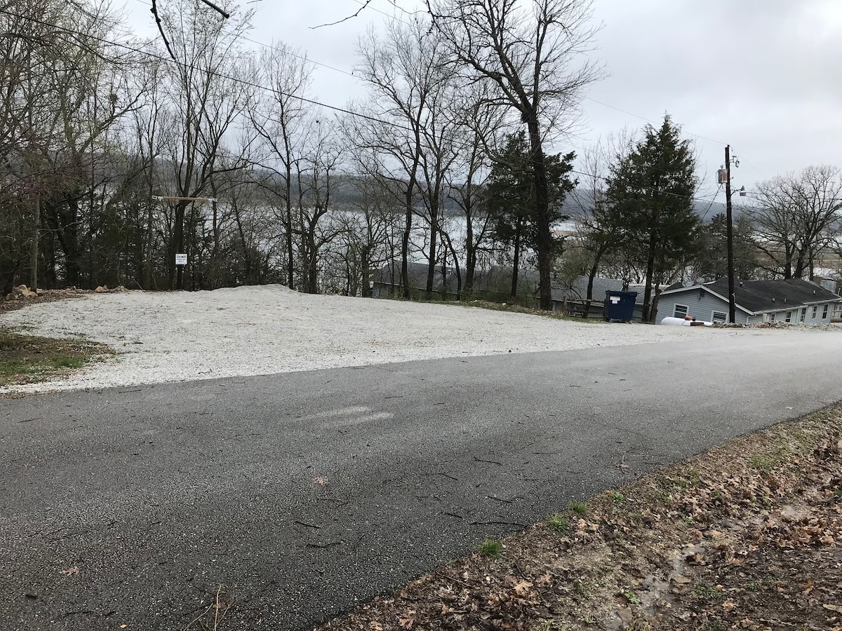 A gravel parking area is seen adjacent to a paved road, with trees lining the space. The view beyond features a lake, partially obscured by branches, with a cloudy sky above. The area illustrates a serene setting conducive for outdoor activities.