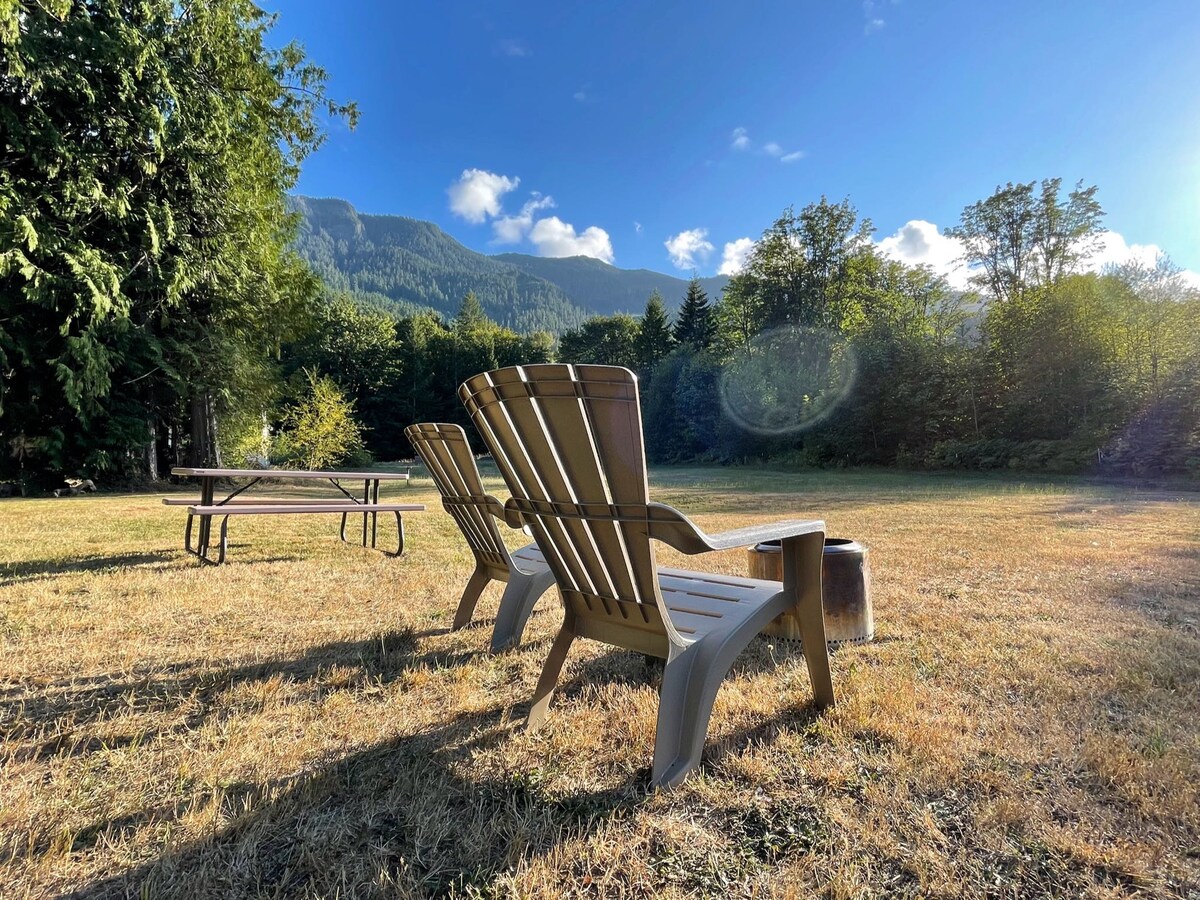 Two wooden Adirondack chairs are positioned on a grassy area, facing a picturesque view of rolling mountains under a clear blue sky. A picnic table and a small fire-pit are situated nearby, enhancing the outdoor seating experience in this peaceful setting.