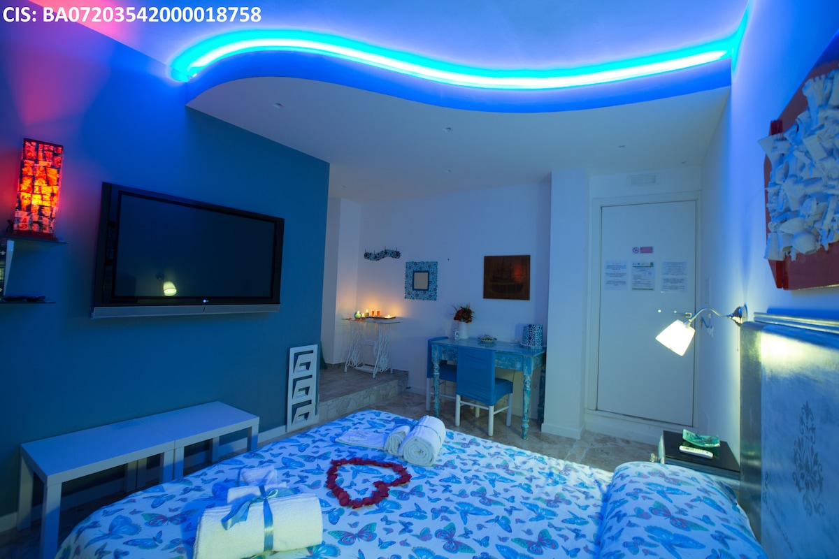 A modern guest room presents soft blue ambient lighting along the ceiling. A spacious bed is adorned with a patterned duvet cover and neatly arranged towels. A flat-screen TV is mounted on the wall, with a small seating area and desk visible in the background.