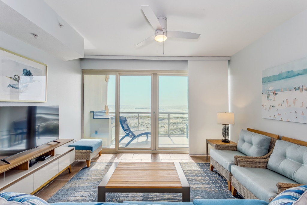 A modern living area features a light blue sofa and matching armchair, arranged around a wooden coffee table. A flat-screen TV sits on a clean-lined media console. Floor-to-ceiling windows offer a view of the balcony and the Gulf beyond, inviting natural light into the space.