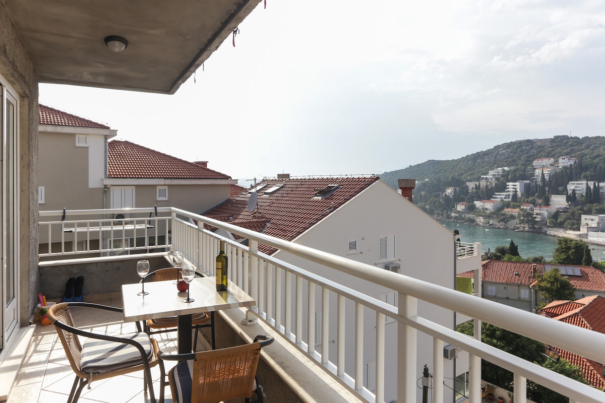 A furnished balcony overlooks the scenic sea view and surrounding rooftops. A dining table is set for two with a bottle of wine and glasses, while two wicker chairs provide comfortable seating. The soft light of day enhances the relaxing atmosphere outside.