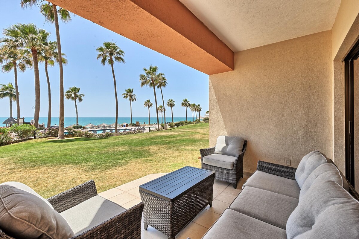 A comfortable outdoor seating area features plush gray cushions on wicker furniture, facing a lush grassy area. Towering palm trees and ocean views are visible beyond the patio, inviting relaxation and light breezes.