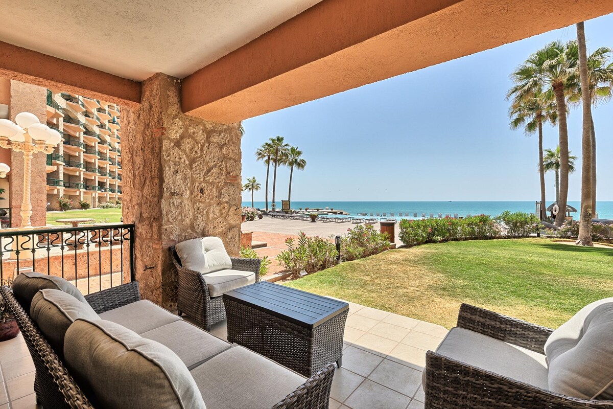 A comfortable outdoor seating area features modern wicker furniture, including a sofa and coffee table. The view is directed towards the ocean and palm trees, with well-maintained grass providing an inviting outdoor space. The tranquility of the environment is enhanced by the distant wave sounds.