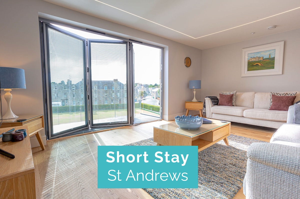A spacious living area is characterized by large bifold doors, allowing natural light to fill the room and providing access to a private balcony. Neutral-toned furniture and decor create a modern aesthetic, complemented by a simple coffee table and an area rug set against a wooden floor.
