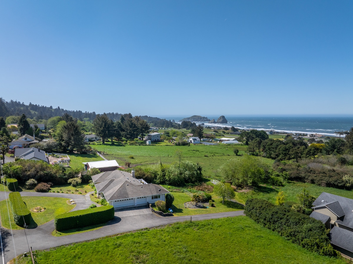 An aerial view captures the spacious property and its surroundings, showcasing lush green fields and a distant ocean view. The coastline features rocky outcrops, while various buildings, including the main home and nearby structures, are positioned harmoniously within the landscape.