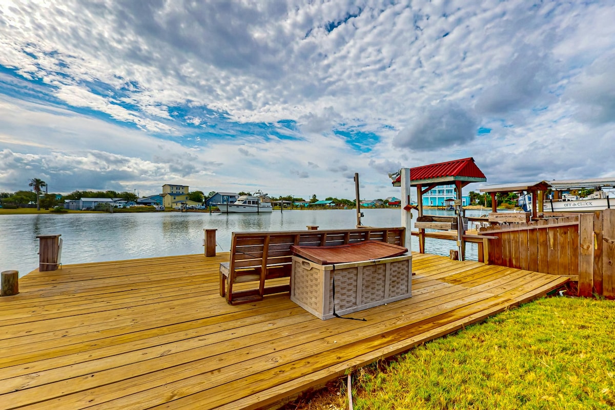 A private dock extends over the water, featuring wooden decking and a seating area equipped with a stylish couch and a coffee table. Calm bay waters reflect the overcast sky, while grassy areas border the dock, enhancing the serene outdoor environment.