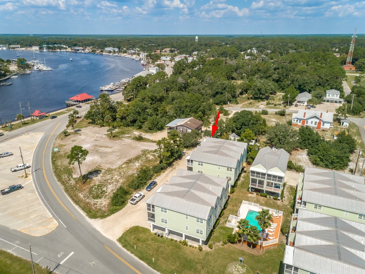 An aerial view captures the property situated near a waterway, surrounded by greenery. Nearby buildings and parking are visible, along with boats docked in the marina. The layout highlights the convenient location, with road access and open space surrounding the structures.