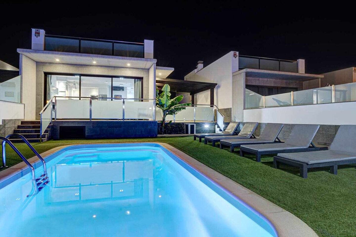 A modern villa is illuminated at night, featuring a private swimming pool surrounded by well-maintained grass. Sun loungers are positioned nearby, and large glass doors lead into a spacious living area, enhancing the connection between indoor and outdoor spaces.