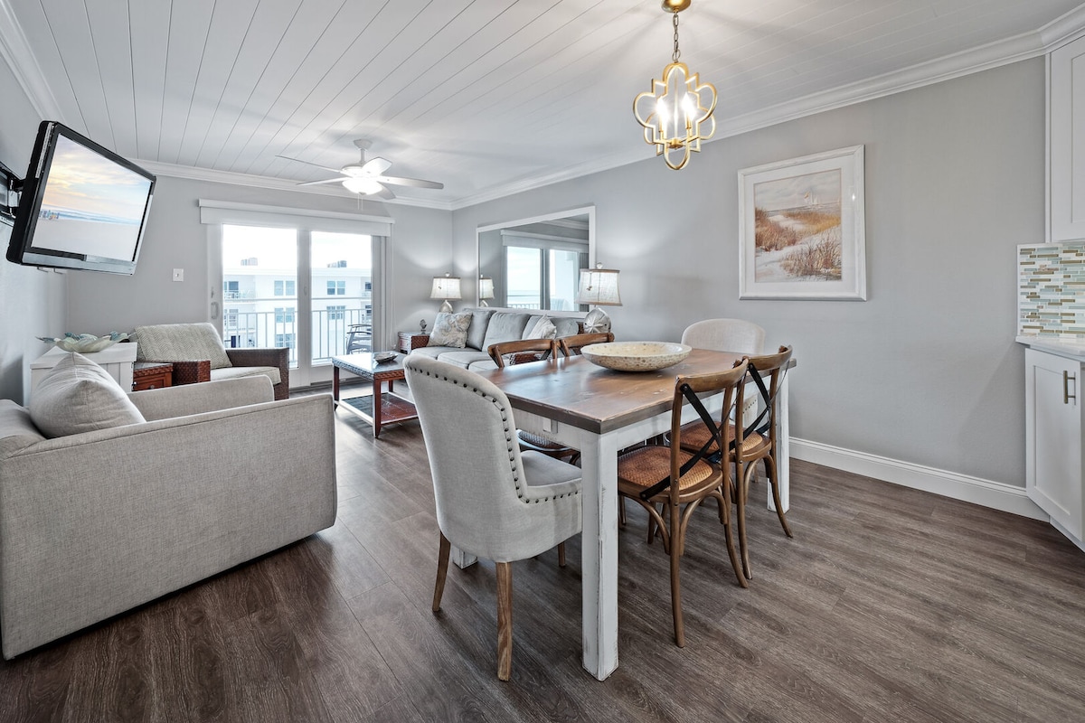 A bright living area is showcased, featuring a comfortable seating arrangement with light-colored sofas and stylish chairs. A dining table with wooden accents is centrally positioned, accompanied by six chairs. The space is enhanced by natural light from large windows offering views of the outdoors.