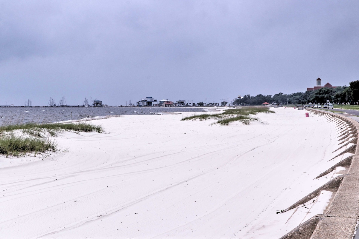 A serene beach scene is captured with soft white sand extending alongside calm waters. Coastal grasses frame the shoreline, while a quiet boardwalk leads toward the distant landscape featuring structures along the shore, creating a tranquil environment.