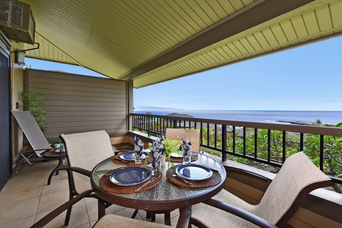 A private furnished lanai features a dining table set for four, showcasing ocean views. Two comfortable lounge chairs are positioned nearby, with greenery in the background and a tranquil sky above. The space is designed for relaxation and enjoyment of the scenic surroundings.