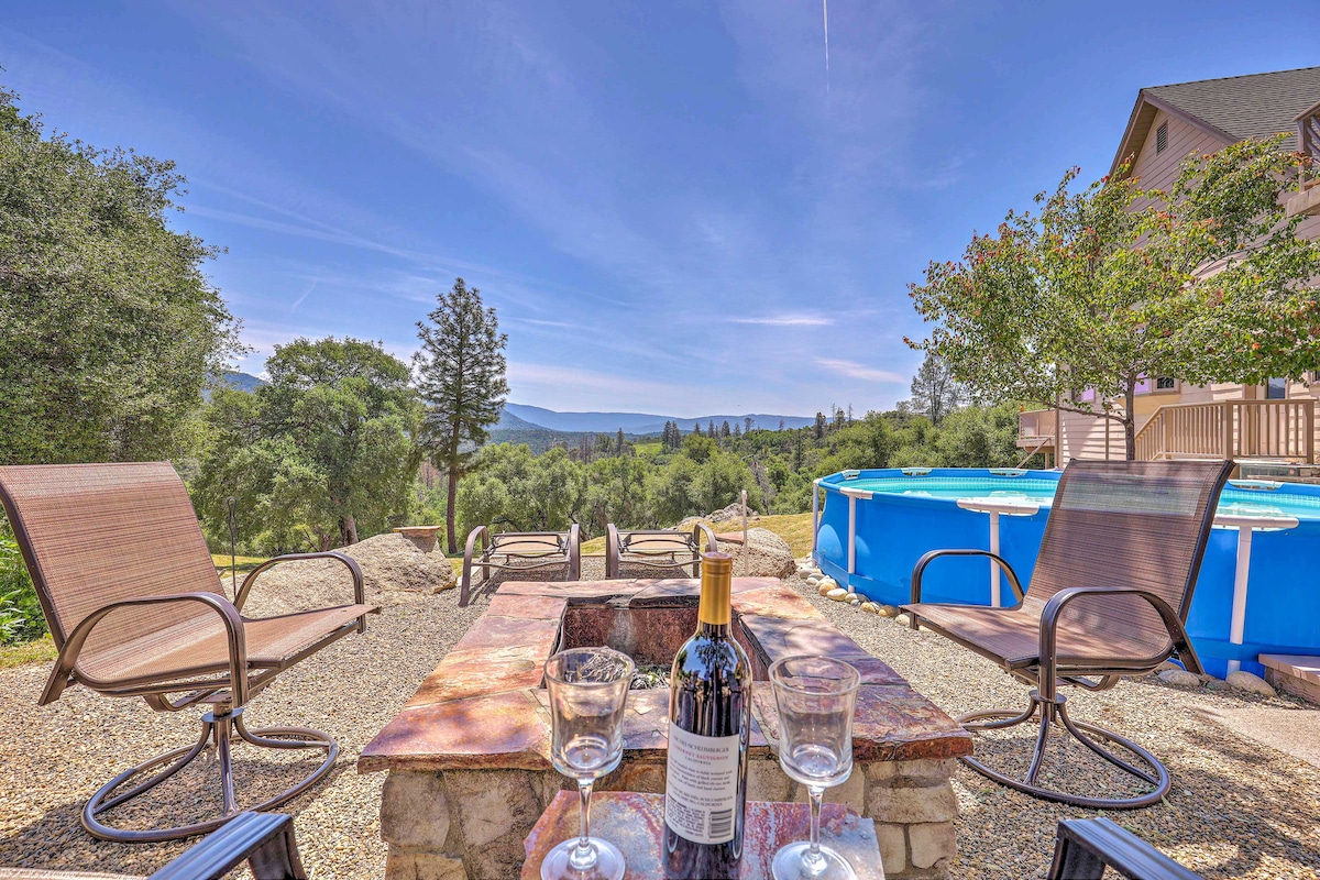 An outdoor seating area features several lounge chairs arranged around a stone fire pit. A bottle of wine and glasses are placed on the table. In the background, a view of lush greenery and soft mountains can be seen under a clear blue sky.