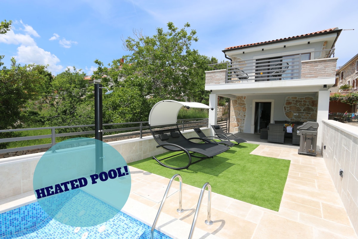 A heated outdoor pool is seen with a light blue mosaic pattern, accompanied by a grassy area. Two lounge chairs provide seating nearby. The house features a balcony with a railing, and plants create a lush environment around the pool area.