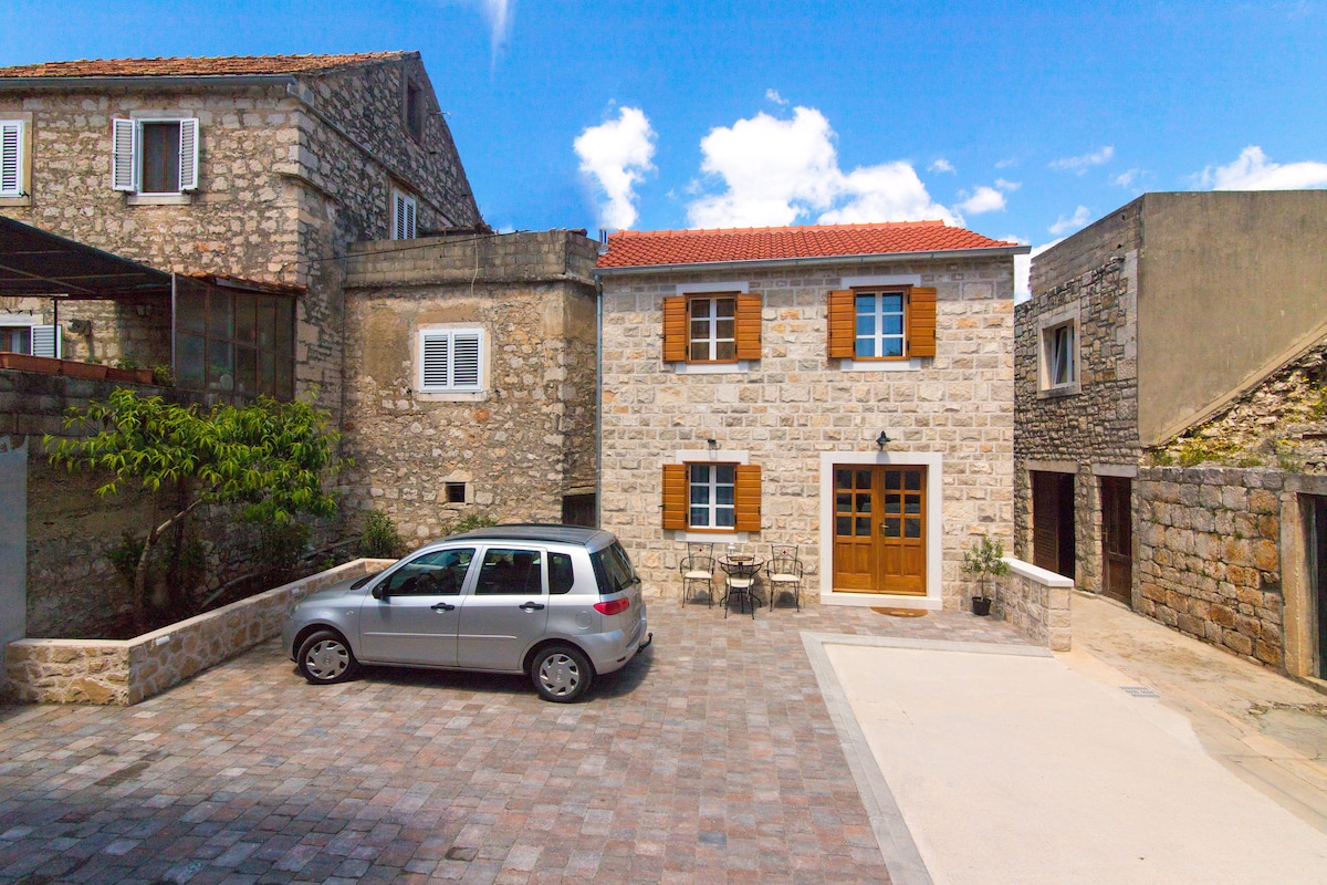 The exterior of a stone house is shown, featuring traditional wooden shutters and a welcoming entryway. A car is parked on the paved courtyard, which includes seating for two beside the entrance. Surrounding buildings display a mix of stone textures under a clear blue sky.