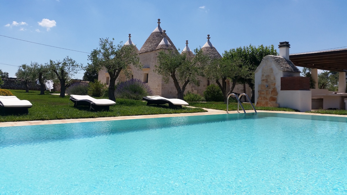 A sparkling pool is framed by a well-maintained lawn and olive trees, with lounge chairs positioned around the edges for relaxation. The iconic trulli buildings are visible in the background, showcasing their unique cone-shaped roofs under a clear blue sky.