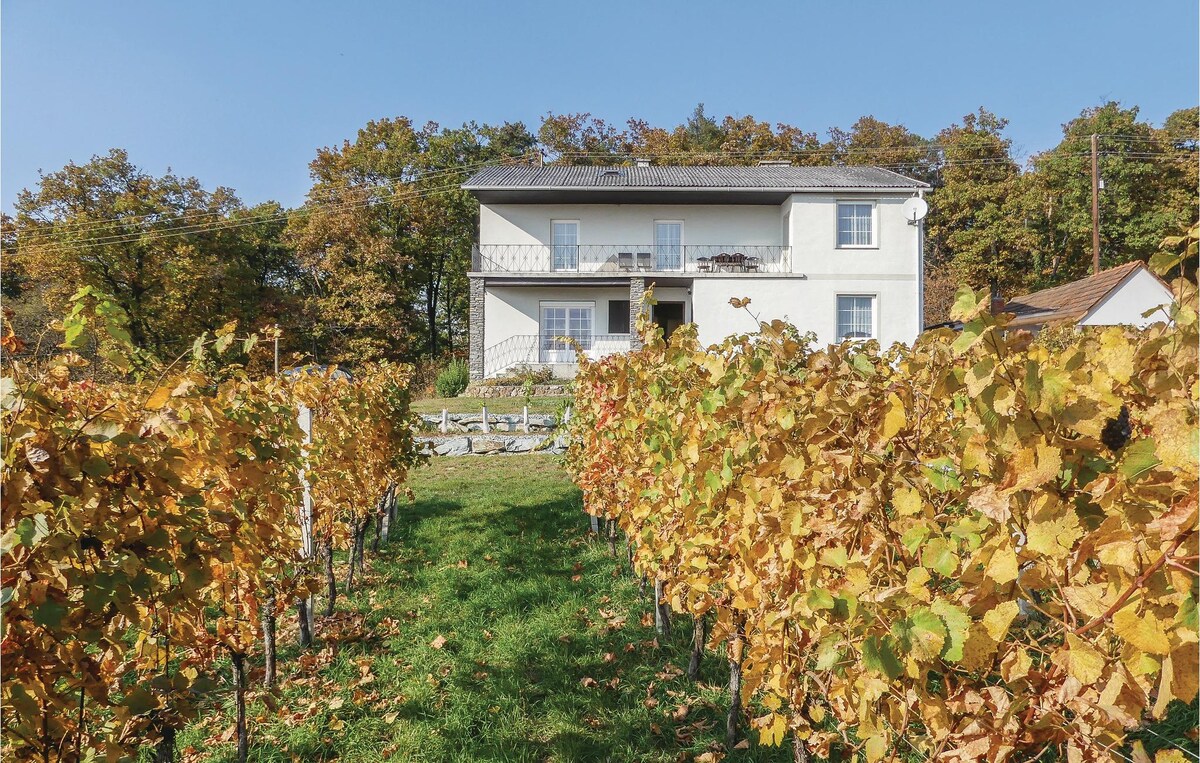 A charming cottage is situated among rows of vibrant, autumn-colored vineyards. The two-story structure features a balcony, and its white exterior contrasts with the lush greenery surrounding it. A clear blue sky enhances the peaceful setting, perfect for nature lovers.