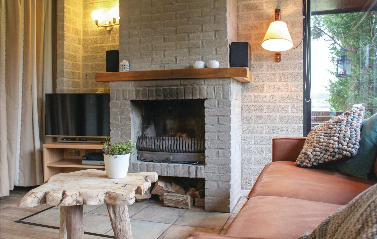 A cozy living area features a rustic fireplace made of light-colored bricks, accompanied by modern speakers. A wooden coffee table complements a comfortable sofa adorned with decorative pillows. Soft lighting is provided by sconces and natural light enters through large windows framed with curtains.