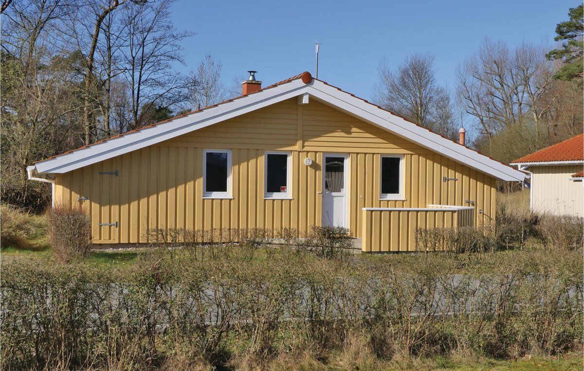 A wooden vacation home is shown, featuring a sloped roof and a warm yellow exterior. Large windows reflect natural light, while a small porch is visible at the front. Surrounding greenery adds a touch of nature to the charming setting.