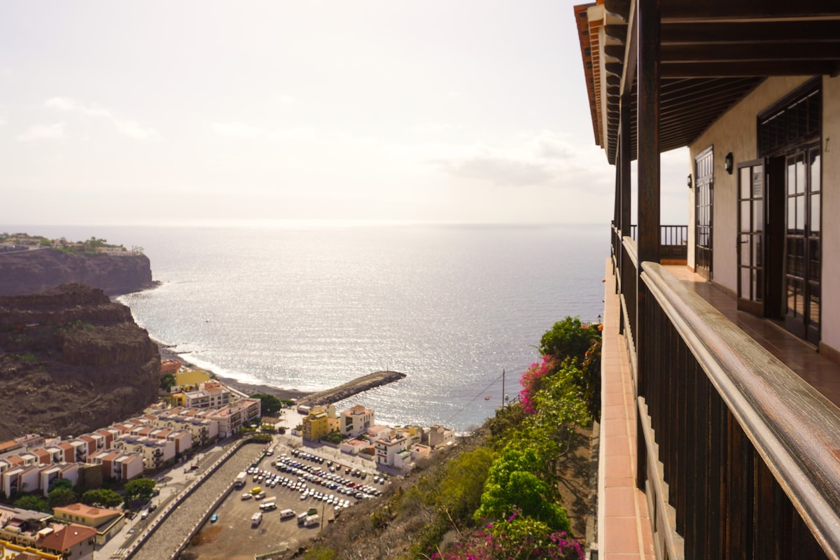 A panoramic view of the coastline and bay is captured from the terrace of the villa. The tranquil sea glistens under sunlight, while colorful buildings line the shore below. Lush greenery frames the scene, enhancing the sense of privacy and tranquility offered by the location.