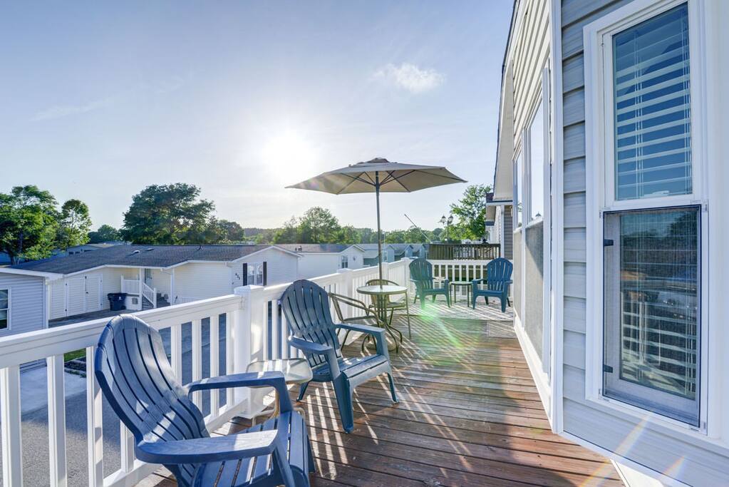 A wrap-around deck is presented, featuring several adirondack chairs arranged for outdoor seating. An umbrella offers shade, while sunlight bathes the area. The view includes neighboring units and greenery, enhancing the casual outdoor atmosphere.