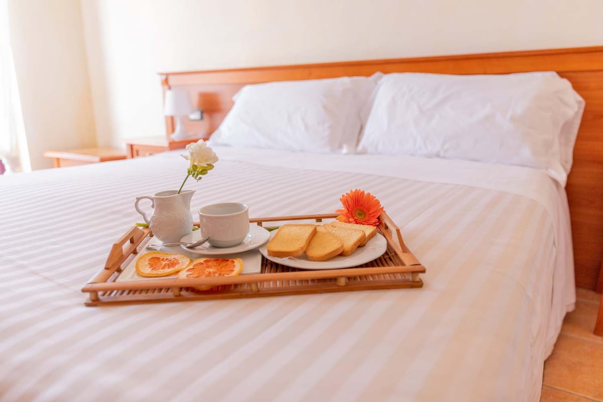 A spacious bed is presented with a tray that holds a small vase with a flower, a cup, and sliced bread with orange. Natural light softly illuminates the room, creating a fresh and inviting setting.