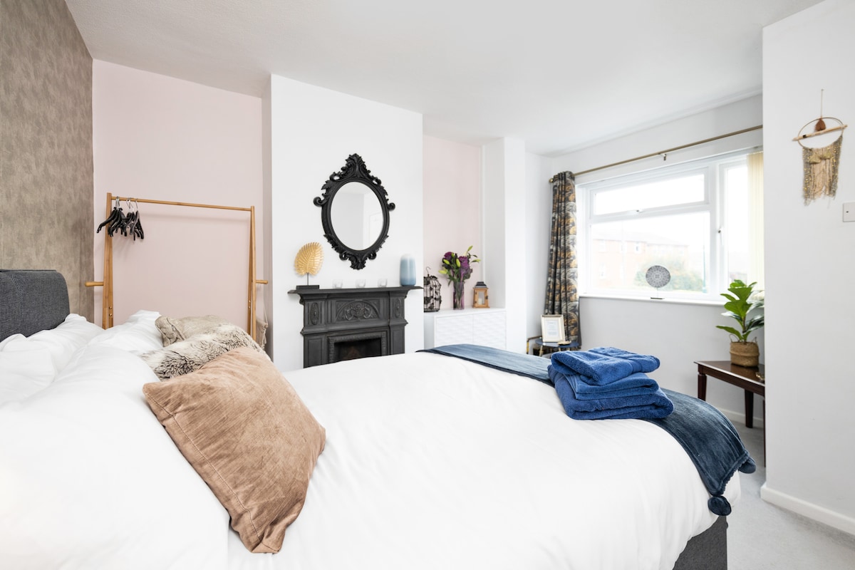 A well-lit bedroom features a spacious bed adorned with decorative cushions and a soft throw. A vintage mirror hangs above a dark fireplace, while a window allows natural light to flow in. Potted plants and decorative elements enhance the inviting atmosphere.