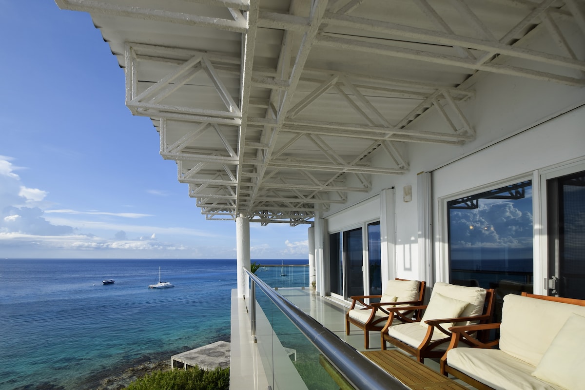 A wraparound balcony features comfortable seating areas, offering direct views of the serene ocean. The expansive space is complemented by a clear glass railing, enhancing the unobstructed sightlines of the turquoise waters and distant sailboats under a bright sky.