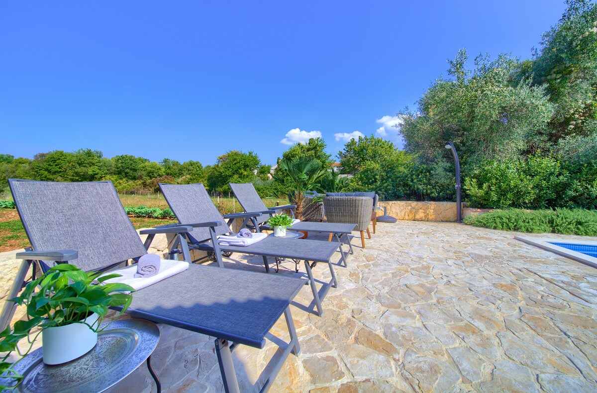 A stone patio area features loungers arranged in a row, complemented by a side table with a potted plant. Lush greenery surrounds the space, providing a natural backdrop. The clear blue sky enhances the serene environment, inviting relaxation near the swimming pool.