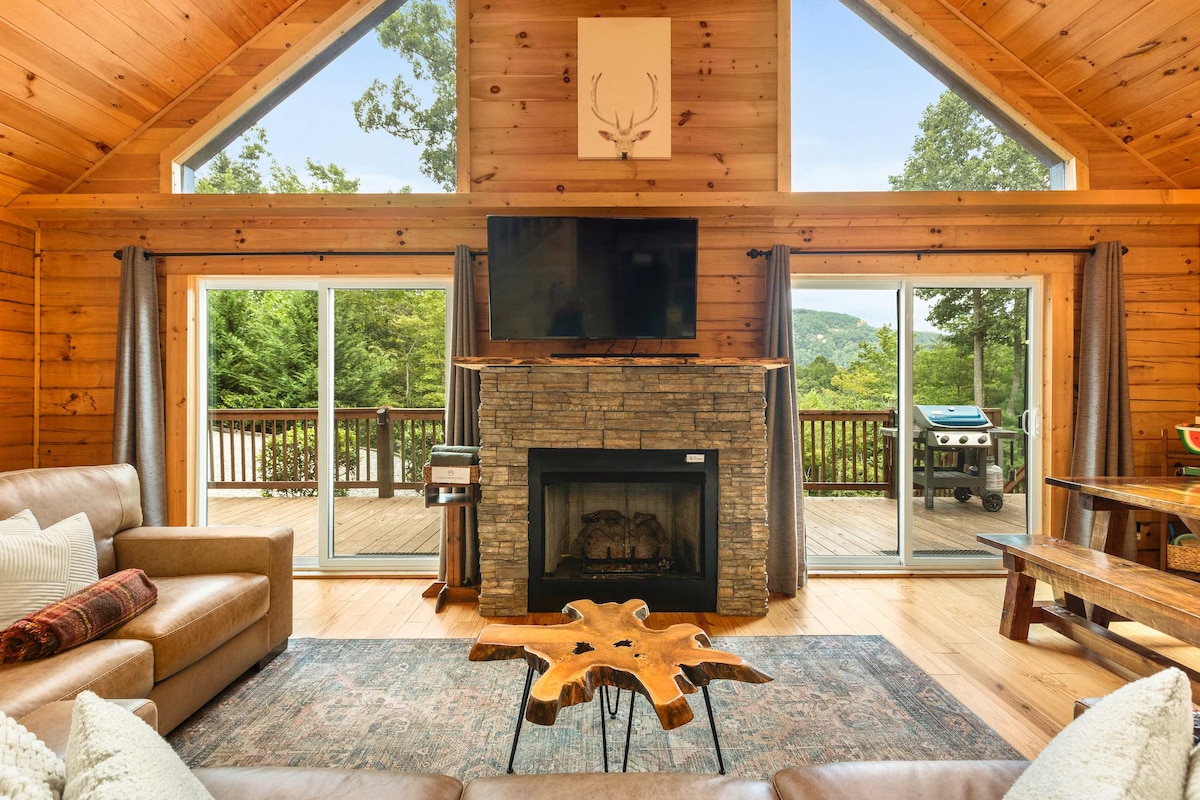 The living area features a cozy leather sofa and matching love seat, positioned around a unique wood coffee table. A natural stone fireplace stands beneath a mounted television, while large sliding glass doors provide access to a spacious deck, framing the mountain views.