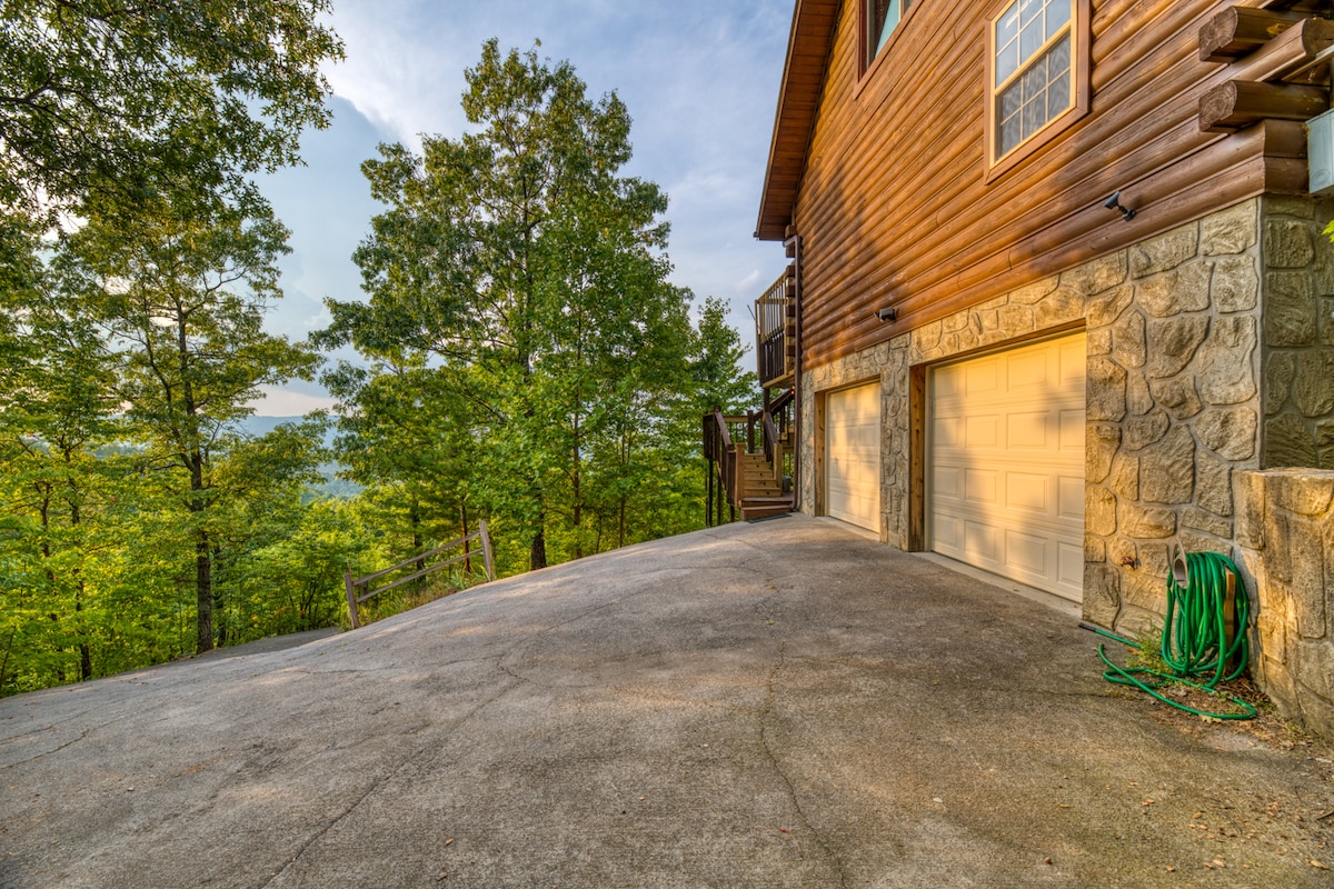 The exterior driveway of the cabin is visible, with a combination of stone and wood textures on the structure. Lush greenery surrounds the area, creating a serene environment. A hose is coiled near the paved surface, while steps lead toward the entrance of the cabin.
