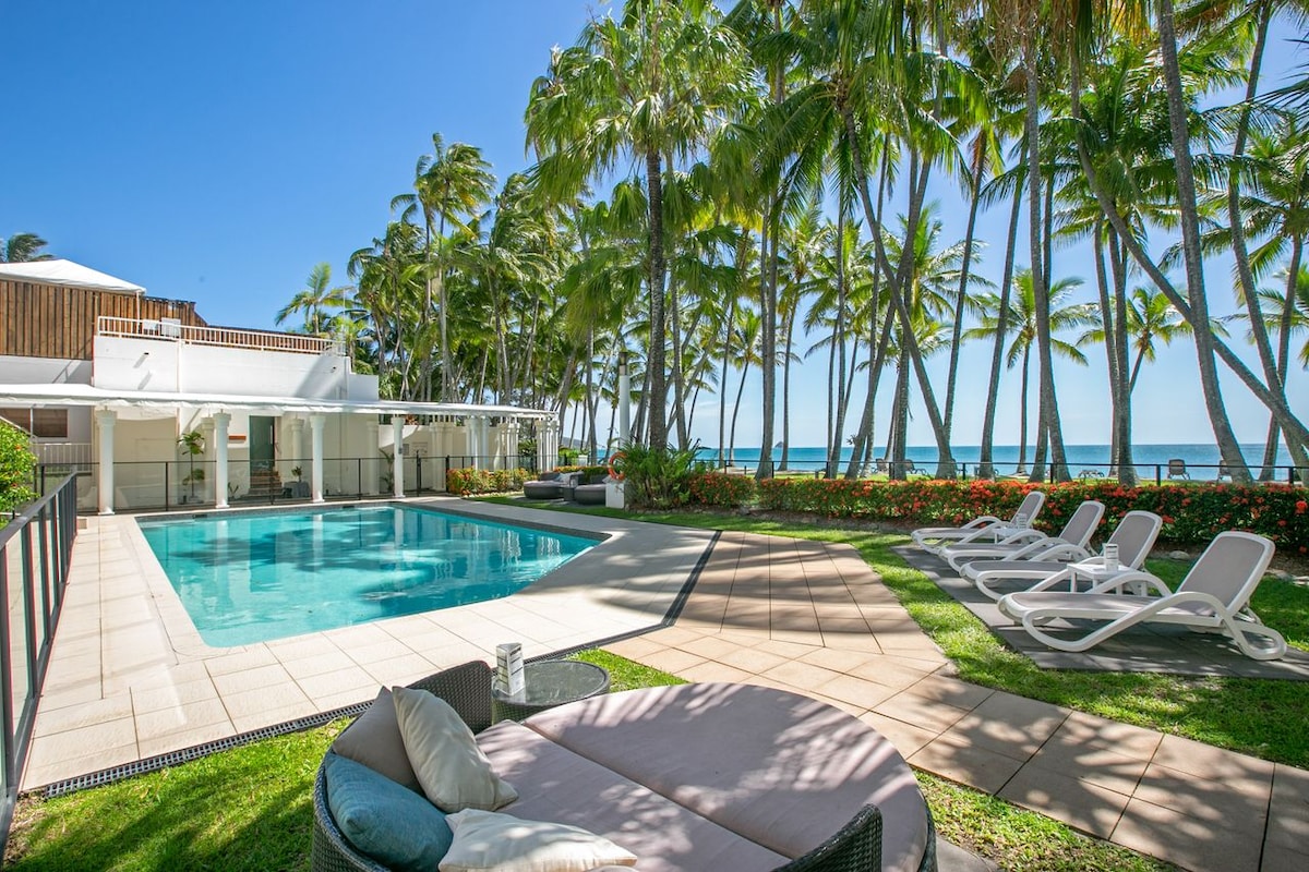 The outdoor area features a tranquil swimming pool surrounded by lush palm trees and landscaped gardens. Comfortable lounge chairs line the poolside, providing ideal spots for relaxation. The ocean is visible in the background, enhancing the serene atmosphere of the space.