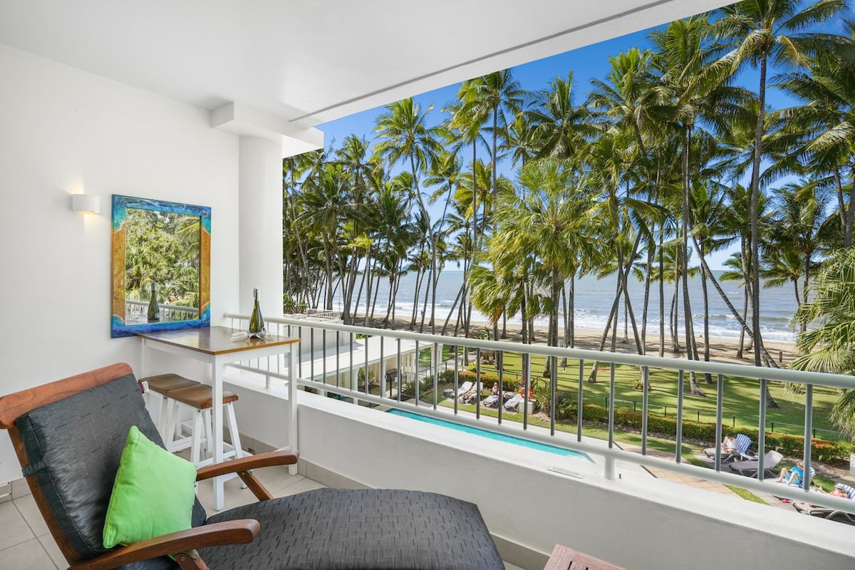 A private balcony features a lounge chair and a small table, offering stunning views of palm trees and the ocean. Sunlight illuminates the outdoor space, enhancing the serene atmosphere of the beachside setting.