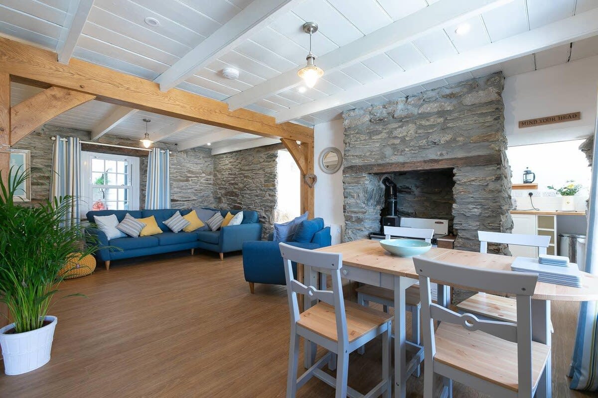 An open-plan living and dining area features exposed wooden beams and a blend of stone and light-colored walls. A blue sectional sofa with colorful cushions is positioned beside a wooden dining table with matching chairs. A wood burner enhances the cozy ambiance, and natural light floods the space.