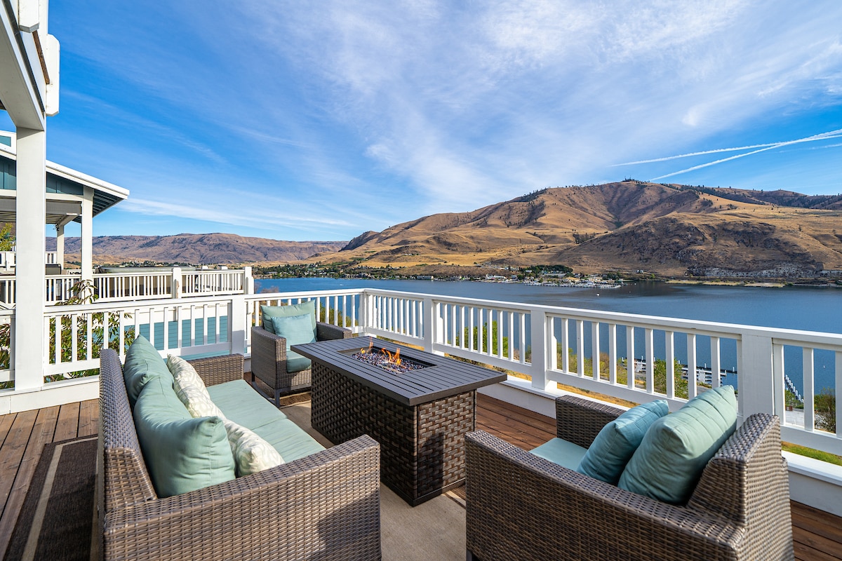 An outdoor seating area features comfortable wicker couches adorned with light blue cushions, positioned around a central fire table. The space offers sweeping views of Lake Chelan and the surrounding hills, with clear skies creating a serene backdrop.