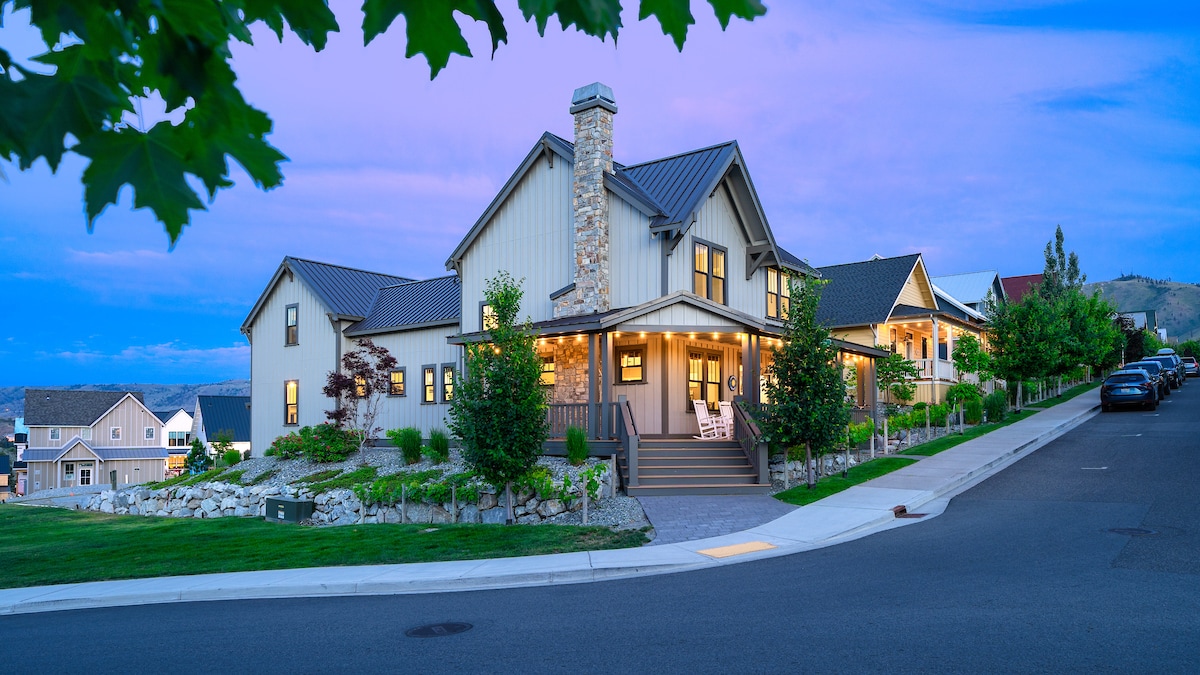 The exterior of a charming lake cottage is illuminated by soft evening lights, showcasing a combination of stone and wood elements. A spacious porch features rocking chairs and a well-manicured yard. The surrounding neighborhood is visible, highlighting a peaceful, welcoming atmosphere.