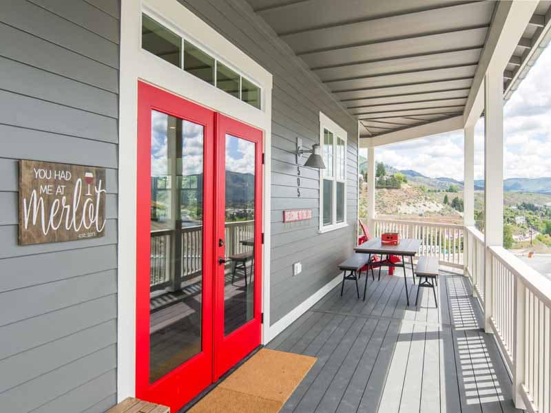A welcoming porch features bold red double doors, contrasting with the gray siding. A wooden table and chairs are arranged for dining, with a striking view of the surrounding landscape visible. Sunlight casts soft shadows, highlighting the spaciousness of the area.