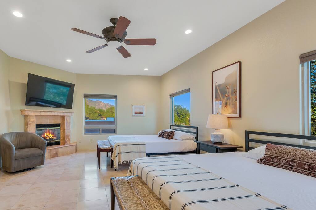 A well-appointed bedroom is featured, showcasing two spacious beds with neutral bedding. Natural light enters through multiple windows, highlighting the fireplace and large television mounted on the wall. A cozy armchair is positioned nearby, creating a comfortable and inviting environment.