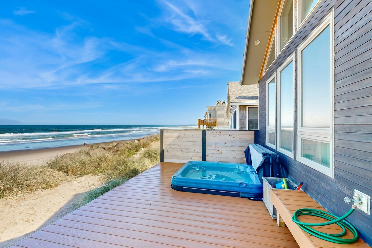 A spacious deck features a private hot tub, providing direct views of the beach and ocean. Soft sands and gentle waves can be seen just steps away. The modern home’s exterior showcases large windows that invite natural light and coastal scenery.