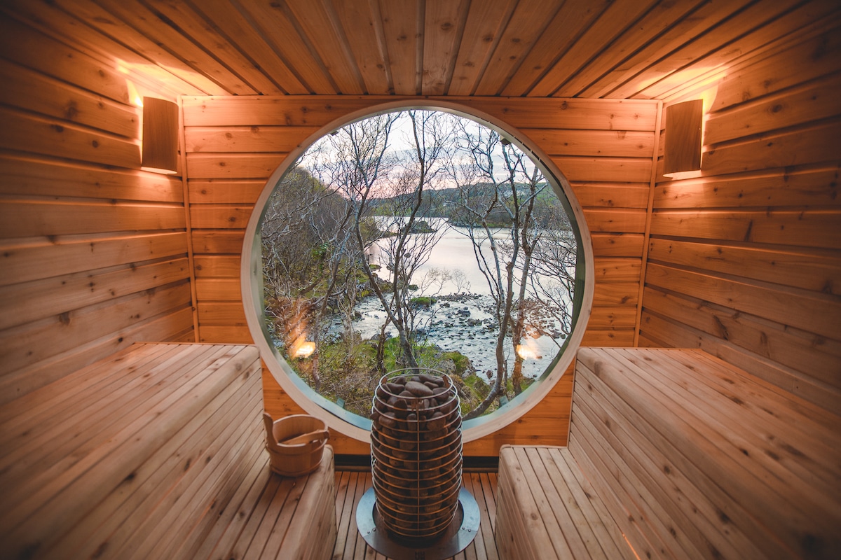 A circular window frames a scenic view of the lake, surrounded by trees. The sauna interior is crafted with warm wooden panels, featuring benches on either side. Soft lighting enhances the tranquil atmosphere, inviting relaxation and a connection with nature.