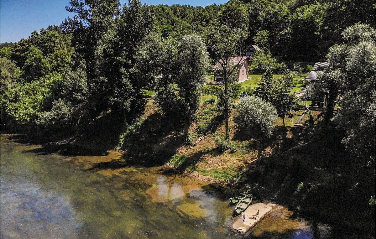 A tranquil view of the cottage perched along the riverbank, surrounded by lush greenery. Two small boats are visible on the water's edge, with a sandy section offering access to the river. The natural landscape creates an inviting escape into nature.