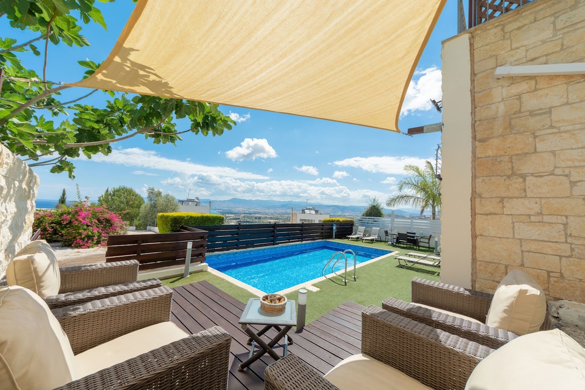 A sunshade provides relief over a cozy seating area featuring wicker chairs. The private swimming pool, framed by lush greenery, is visible in the background along with a spacious outdoor decking area. Panoramic views of the surrounding landscape are captured under a clear blue sky.