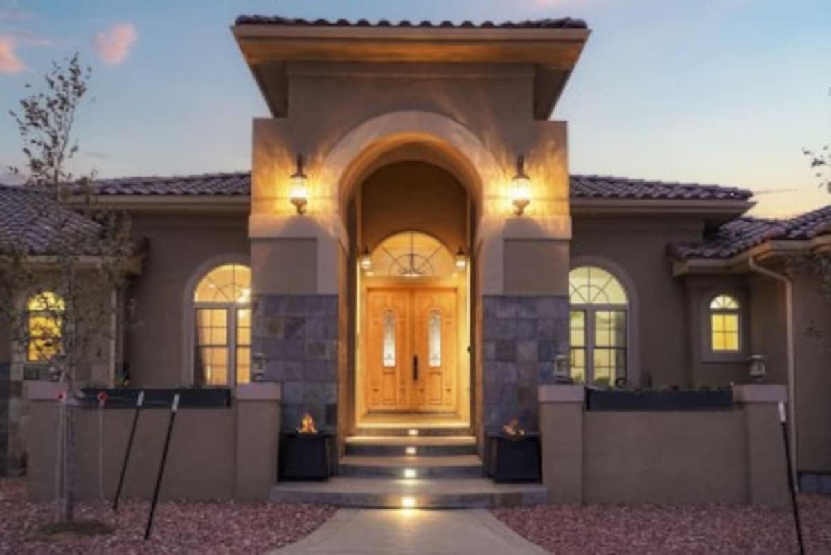 The exterior entrance of the mansion showcases an elegant double door flanked by two large arched windows. Soft lighting highlights the architectural details, while a stone walkway guides visitors toward the inviting entrance, surrounded by landscaped gardens.