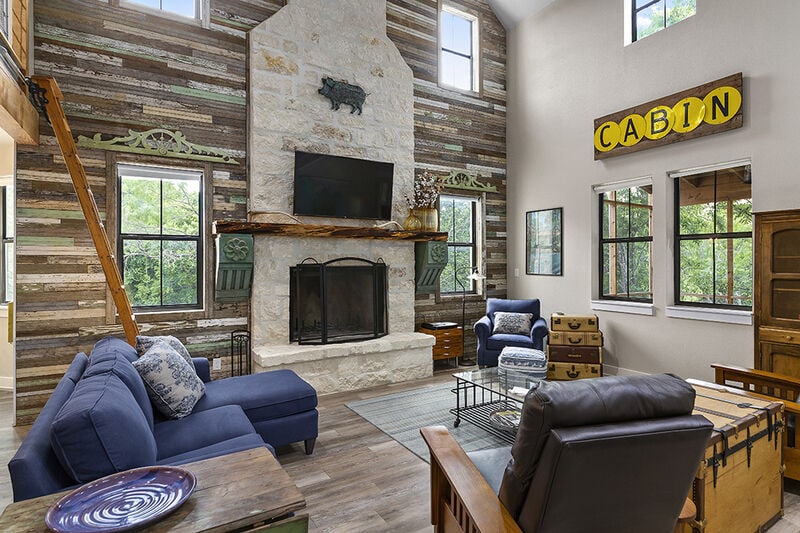 The living room features a stone fireplace, framed by wood paneling and two large windows allowing natural light. Comfortable seating arrangements include a blue sofa and leather chairs, complemented by a rustic coffee table. A sign reading 'CABIN' adds a charming touch to the space.