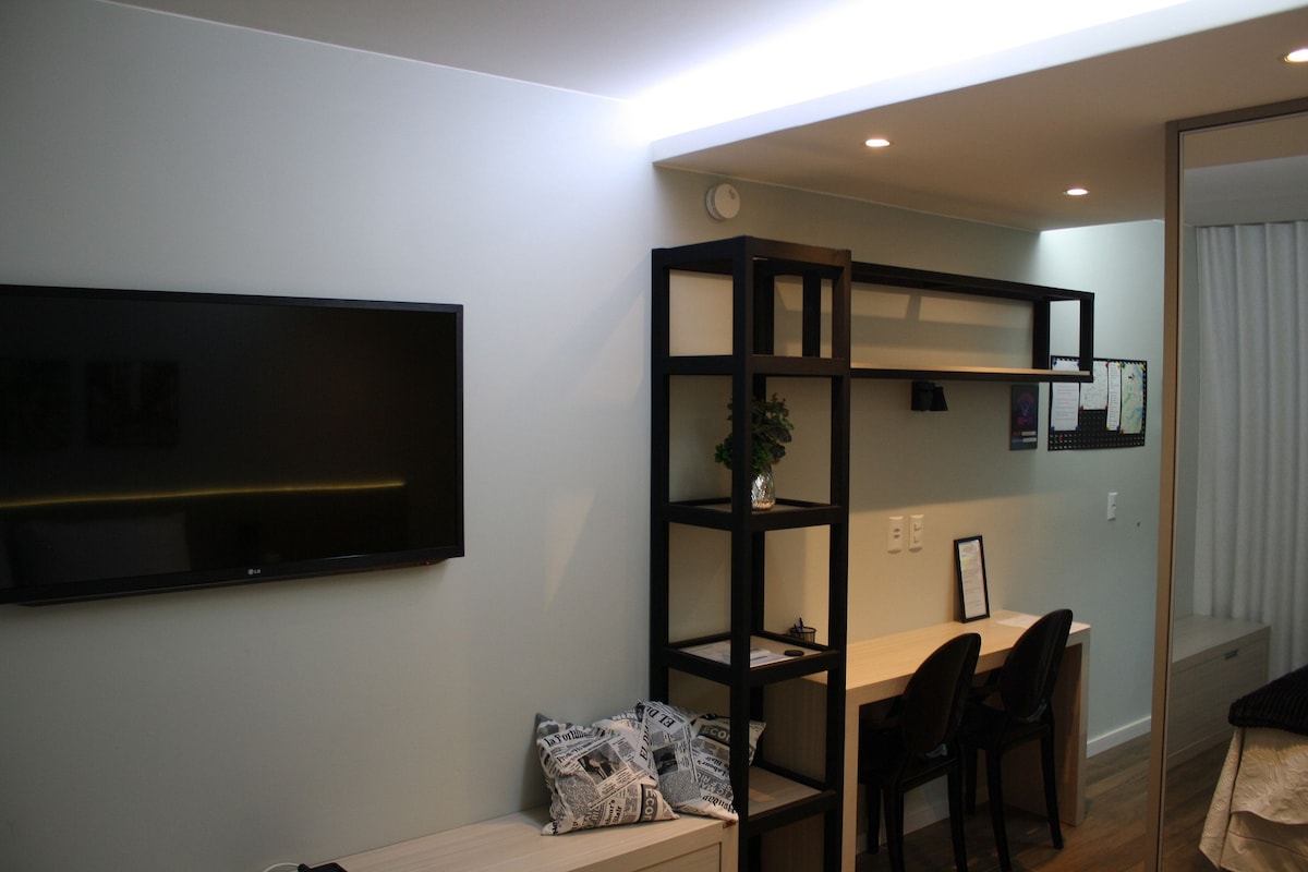 A modern living space features a mounted flat-screen TV above a sleek media console. A minimalist desk with two black chairs is positioned near a decorative bookshelf, which holds a small plant and framed items, enhancing the functional layout.