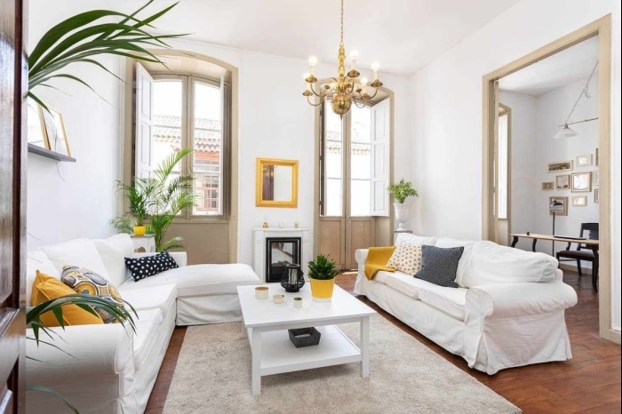 A bright living room features large windows that provide abundant natural light. Two cozy sofas, adorned with decorative cushions, face a central coffee table. A small plant adds a touch of greenery, while a stylish chandelier hangs from the ceiling, contribute to the room's inviting atmosphere.