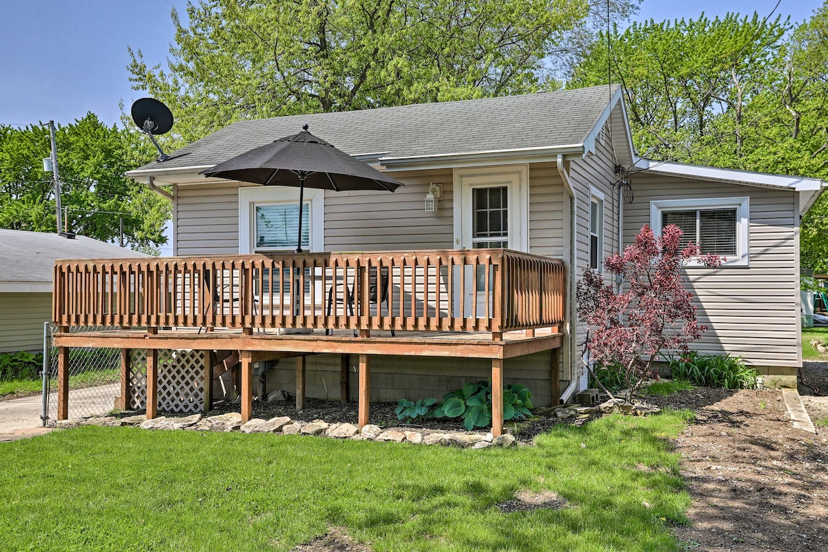 A single-story home with a light-colored exterior is showcased. A wooden deck is furnished with an umbrella and seating, surrounded by a green lawn and garden. Trees and foliage provide a natural backdrop, creating a relaxed outdoor environment.