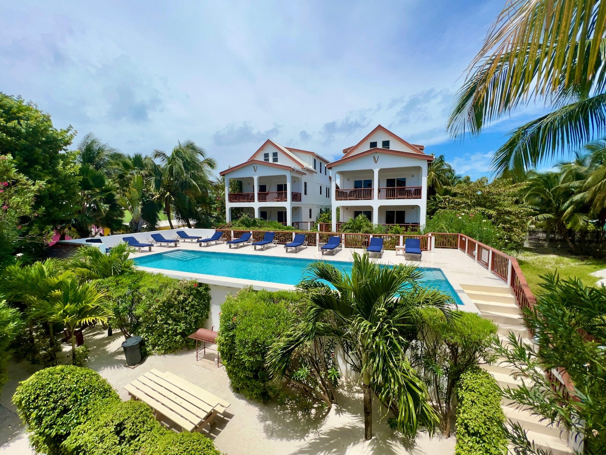 A spacious pool area is surrounded by tropical greenery and sun loungers with padded cushions. The three-story building features multiple balconies overlooking the pool, promoting a relaxed and inviting environment for relaxation and leisure.